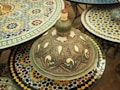 An intricately designed decorative Moroccan tagine lid rests on a colorful mosaic table. The lid is adorned with detailed geometric patterns and an array of colors, including green, blue, and red accents against a metallic and ivory background. Surrounding the tagine are several other tables covered with similarly vibrant mosaic patterns.