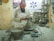 A potter is focused on shaping a clay vessel on a pottery wheel in a workshop filled with clay sculptures and tools. The workspace is cluttered with clay materials, and the wall is adorned with posters and a FedEx advertisement.