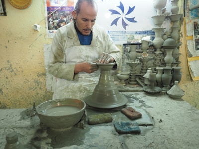 A potter is focused on shaping a clay vessel on a pottery wheel in a workshop filled with clay sculptures and tools. The workspace is cluttered with clay materials, and the wall is adorned with posters and a FedEx advertisement.