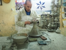 A potter is focused on shaping a clay vessel on a pottery wheel in a workshop filled with clay sculptures and tools. The workspace is cluttered with clay materials, and the wall is adorned with posters and a FedEx advertisement.