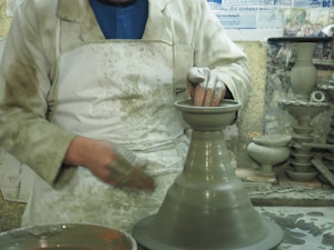 A person is engaged in pottery making, wearing a white apron and shaping clay on a spinning wheel. The workspace is cluttered with pottery tools and finished clay pieces in the background, suggesting an active studio setting.