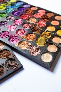 A variety of colorful powders are arranged in a palette, featuring shades like pink, purple, yellow, and blue. Some sections show the powders crushed and spilled out, creating a textured appearance. The palette is placed on a white surface.