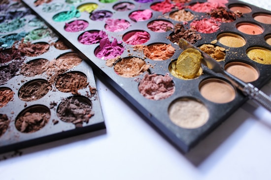 A collection of colorful eyeshadow palettes is displayed, featuring various shades including blues, greens, pinks, purples, browns, and yellows. Some of the makeup is crushed and scattered, showing texture and creating a sense of used and creative disarray. A metal tool is placed on one of the palettes, suggesting an application or mixing in progress.