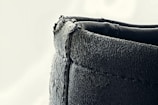A close-up view of a textured, dark-colored leather edge with visible stitching. The surface appears slightly worn and rugged, with some frayed edges on the top side.