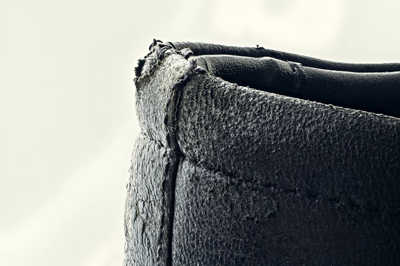A close-up view of a textured, dark-colored leather edge with visible stitching. The surface appears slightly worn and rugged, with some frayed edges on the top side.