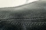 Detailed close-up of black leather material with visible stitching along the seams. The texture appears smooth with slight signs of wear and creasing.
