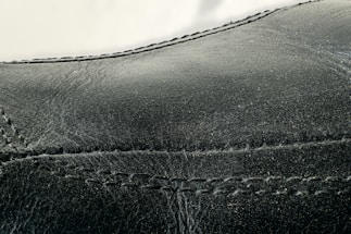 Detailed close-up of black leather material with visible stitching along the seams. The texture appears smooth with slight signs of wear and creasing.