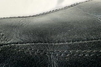 Detailed close-up of black leather material with visible stitching along the seams. The texture appears smooth with slight signs of wear and creasing.