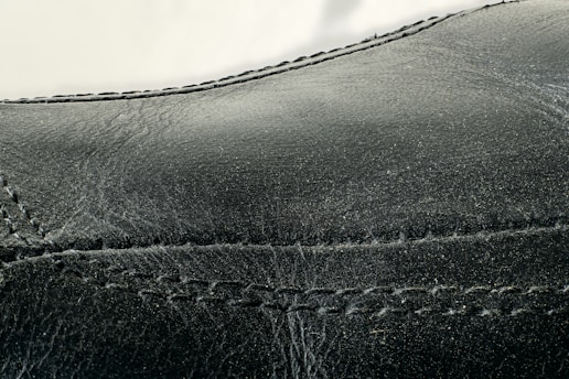Detailed close-up of black leather material with visible stitching along the seams. The texture appears smooth with slight signs of wear and creasing.