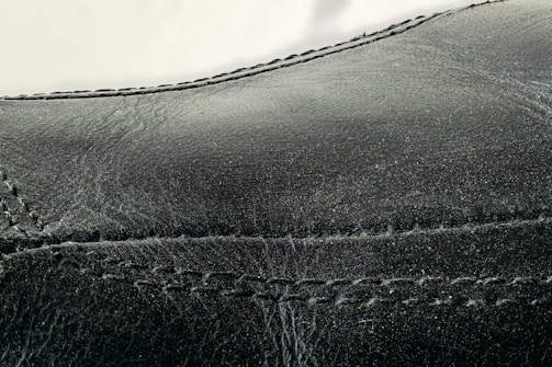 Detailed close-up of black leather material with visible stitching along the seams. The texture appears smooth with slight signs of wear and creasing.