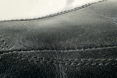 Detailed close-up of black leather material with visible stitching along the seams. The texture appears smooth with slight signs of wear and creasing.