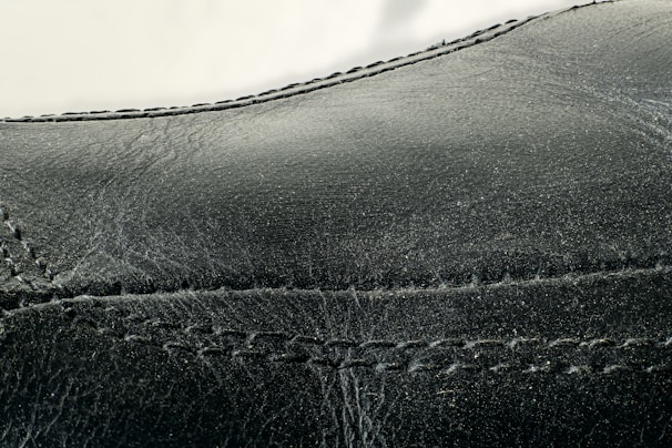 Detailed close-up of black leather material with visible stitching along the seams. The texture appears smooth with slight signs of wear and creasing.