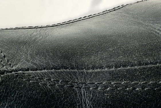 Detailed close-up of black leather material with visible stitching along the seams. The texture appears smooth with slight signs of wear and creasing.