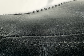Detailed close-up of black leather material with visible stitching along the seams. The texture appears smooth with slight signs of wear and creasing.