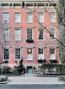 A classic urban townhouse with a pink brick facade is visible, featuring multiple windows with white frames and decorative moldings. An iron fence surrounds the small front garden, which is adorned with shrubbery and trees. The street in front appears quiet and is lined with a sidewalk.