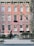 A classic urban townhouse with a pink brick facade is visible, featuring multiple windows with white frames and decorative moldings. An iron fence surrounds the small front garden, which is adorned with shrubbery and trees. The street in front appears quiet and is lined with a sidewalk.