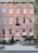 A classic urban townhouse with a pink brick facade is visible, featuring multiple windows with white frames and decorative moldings. An iron fence surrounds the small front garden, which is adorned with shrubbery and trees. The street in front appears quiet and is lined with a sidewalk.