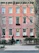 A classic urban townhouse with a pink brick facade is visible, featuring multiple windows with white frames and decorative moldings. An iron fence surrounds the small front garden, which is adorned with shrubbery and trees. The street in front appears quiet and is lined with a sidewalk.