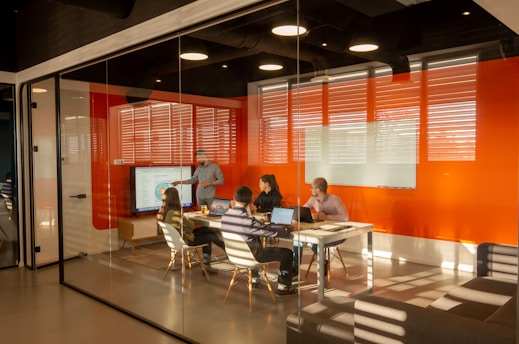 A conference room with glass walls contains a group of people seated around a table, some with laptops. One individual is standing and pointing at a large screen displaying a colorful diagram. Bright orange walls and ample lighting contribute to the modern aesthetic.