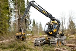 A modern forestry machine with a long articulated arm is actively cutting and processing trees in a forested area. It has large, robust wheels adapted for rough terrain and is surrounded by felled trees and fresh logs.