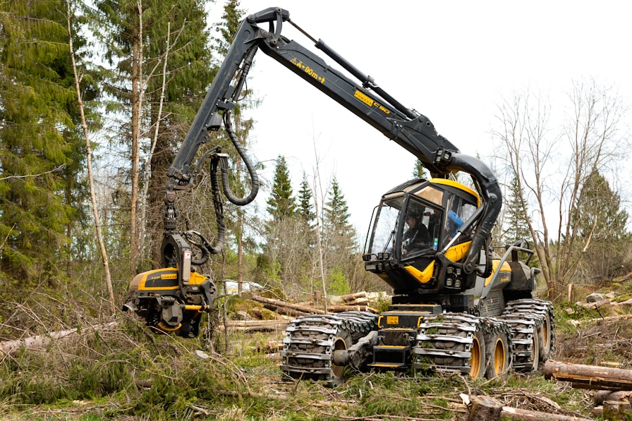 A modern forestry machine with a long articulated arm is actively cutting and processing trees in a forested area. It has large, robust wheels adapted for rough terrain and is surrounded by felled trees and fresh logs.
