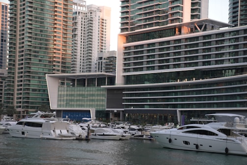 A marina filled with numerous white yachts and boats docked on calm water in front of modern high-rise buildings. The buildings feature a sleek, contemporary design with large glass windows and multiple balconies. The area appears to be a bustling urban setting with a focus on luxury and waterfront living.