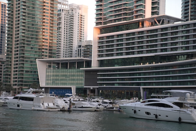 A marina filled with numerous white yachts and boats docked on calm water in front of modern high-rise buildings. The buildings feature a sleek, contemporary design with large glass windows and multiple balconies. The area appears to be a bustling urban setting with a focus on luxury and waterfront living.