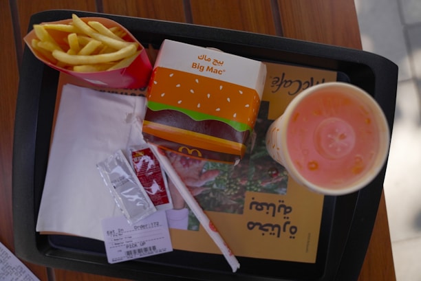 A fast-food meal on a black tray includes a box of Big Mac, a container of fries, a soft drink cup with a lid and straw, two condiment packets, a napkin, and a receipt. The Big Mac packaging is orange with sesame seed illustrations and Arabic script. The fries are in a red container, and the drink appears to be light orange in color.