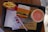 A fast-food meal on a black tray includes a box of Big Mac, a container of fries, a soft drink cup with a lid and straw, two condiment packets, a napkin, and a receipt. The Big Mac packaging is orange with sesame seed illustrations and Arabic script. The fries are in a red container, and the drink appears to be light orange in color.