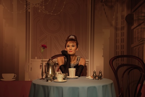A woman dressed elegantly in a black dress and pearl necklace sits at a round table set with fine china and a single red rose. The background features abstract designs reminiscent of a sophisticated cafe setting.