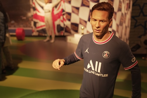 A wax statue dressed in a Paris Saint-Germain football jersey stands indoors. The background includes a checkered flag pattern and a large British flag. Another wax figure in a white racing suit can be seen in the background.