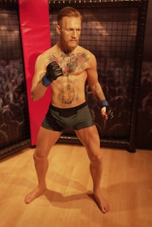 A muscular man with tattoos stands in a fighting pose inside a ring. He is wearing black shorts and has blue wrist wraps. The setting appears to be an arena with a crowd visible in the background.