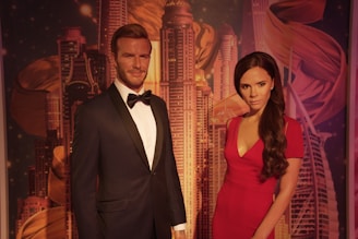 Two life-sized wax figures of a man and a woman dressed in formal attire; the man is wearing a black tuxedo with a bow tie, and the woman is wearing a red gown. They stand side by side, set against a backdrop of a city skyline with tall skyscrapers and abstract artistic elements.