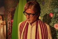 A wax figure of a man wearing a traditional embroidered cream-colored robe with a red and white striped scarf. He has black glasses and gray hair. The background features a green leafy wall adorned with colorful flowers.
