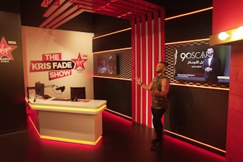 A radio studio setup with modern decor. The room features a desk with a microphone, headphones, and computer monitor. Bright red and white lighting creates an energetic atmosphere. A man stands to the right of the image, gesturing as if speaking to an audience. A sign for 'The Kris Fade Show' is prominently displayed, and a screen shows an advertisement for the 90th Oscars.