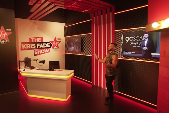 A radio studio setup with modern decor. The room features a desk with a microphone, headphones, and computer monitor. Bright red and white lighting creates an energetic atmosphere. A man stands to the right of the image, gesturing as if speaking to an audience. A sign for 'The Kris Fade Show' is prominently displayed, and a screen shows an advertisement for the 90th Oscars.