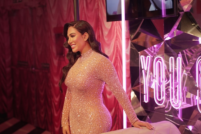 A smiling woman with long dark hair is wearing a glittering, form-fitting dress. She is standing in a room with pink, shimmering curtains and a reflective wall featuring pink neon letters. The ambiance is elegant and glamorous.