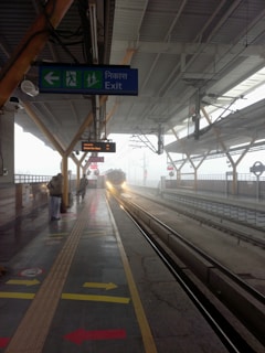 A train station platform with a train approaching on the tracks. There are a few people standing on the platform, and various signs including an exit sign in multiple languages. The platform has directional arrows on the ground, and the scene is enveloped in a misty atmosphere.