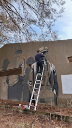 A person is on a ladder painting a mural on a large exterior wall. The mural features a detailed, monochromatic image of three people wearing hats, with one prominently in the foreground. The setting appears to be outdoors with trees visible in the background.