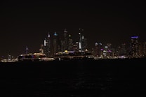 A nighttime cityscape with illuminated skyscrapers and buildings along a coast. The city lights create a vibrant display against the dark sky, with reflections visible on the water. Cruise ships with colorful lights are seen in the foreground, adding to the lively ambiance.