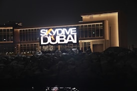A large illuminated sign displaying 'SkyDive Dubai' is prominently featured against a modern building at night. The letters are bright and the sign includes an image of a skydiver. The building has large glass windows and a well-lit facade. In the foreground, there are dark rocks or a rocky terrain leading up to the structure.