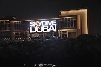 A large illuminated sign displaying 'SkyDive Dubai' is prominently featured against a modern building at night. The letters are bright and the sign includes an image of a skydiver. The building has large glass windows and a well-lit facade. In the foreground, there are dark rocks or a rocky terrain leading up to the structure.