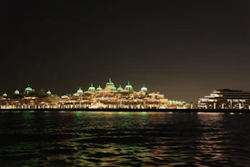 A waterfront view of an illuminated hotel with domed roofs glowing green in a nighttime setting. The building is reflected across the dark water, adding to the tranquil atmosphere.