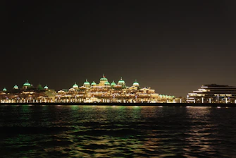 A waterfront view of an illuminated hotel with domed roofs glowing green in a nighttime setting. The building is reflected across the dark water, adding to the tranquil atmosphere.