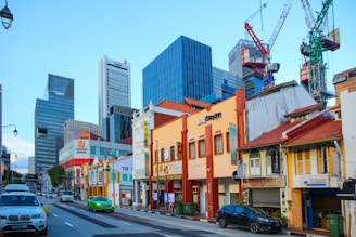 A bustling urban street scene features a mix of traditional low-rise shophouses with colorful facades and tall modern skyscrapers in the background. Several cars, including a green taxi and a silver sedan, are driving along the road. Construction cranes are visible against the blue sky, indicating ongoing development. Business signs adorn the shophouses, and a variety of architectural styles are evident.
