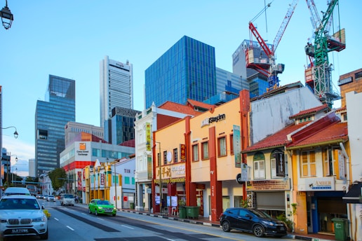 A bustling urban street scene features a mix of traditional low-rise shophouses with colorful facades and tall modern skyscrapers in the background. Several cars, including a green taxi and a silver sedan, are driving along the road. Construction cranes are visible against the blue sky, indicating ongoing development. Business signs adorn the shophouses, and a variety of architectural styles are evident.