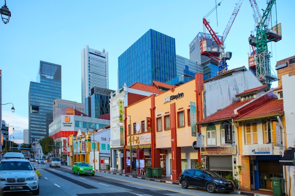 A bustling urban street scene features a mix of traditional low-rise shophouses with colorful facades and tall modern skyscrapers in the background. Several cars, including a green taxi and a silver sedan, are driving along the road. Construction cranes are visible against the blue sky, indicating ongoing development. Business signs adorn the shophouses, and a variety of architectural styles are evident.