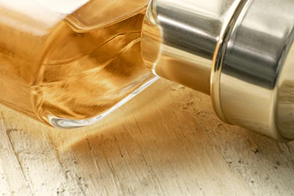 Close-up of a glass bottle filled with golden mustard oil surrounded by mustard seeds on a wooden surface.