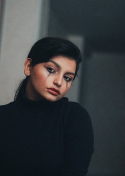 A woman with dramatic makeup, including bold eyeliner and tears drawn from her eyes, is gazing thoughtfully. She wears a dark turtleneck and has a somber expression on her face. The background is muted and indistinct, adding to the overall moody atmosphere.