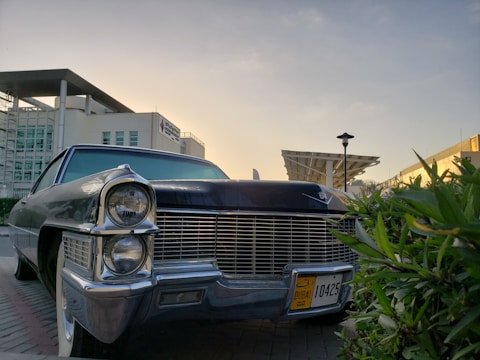 A classic car with an iconic front grille and large headlights is parked on a street. The car has a Dubai license plate and is surrounded by modern architecture, including a building with glass windows and a distinctive overhanging roof. Green foliage is visible in the foreground, and the sky is clear with warm lighting, suggesting a sunset or sunrise.